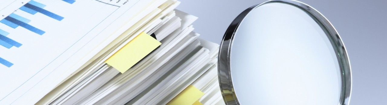 Stack of paper reports with a magnifying glass inspecting them