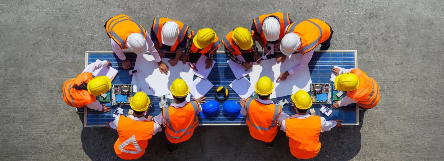 Work site meeting with many people wearing hi-vis and hard hats, gathered around a table outdoors
